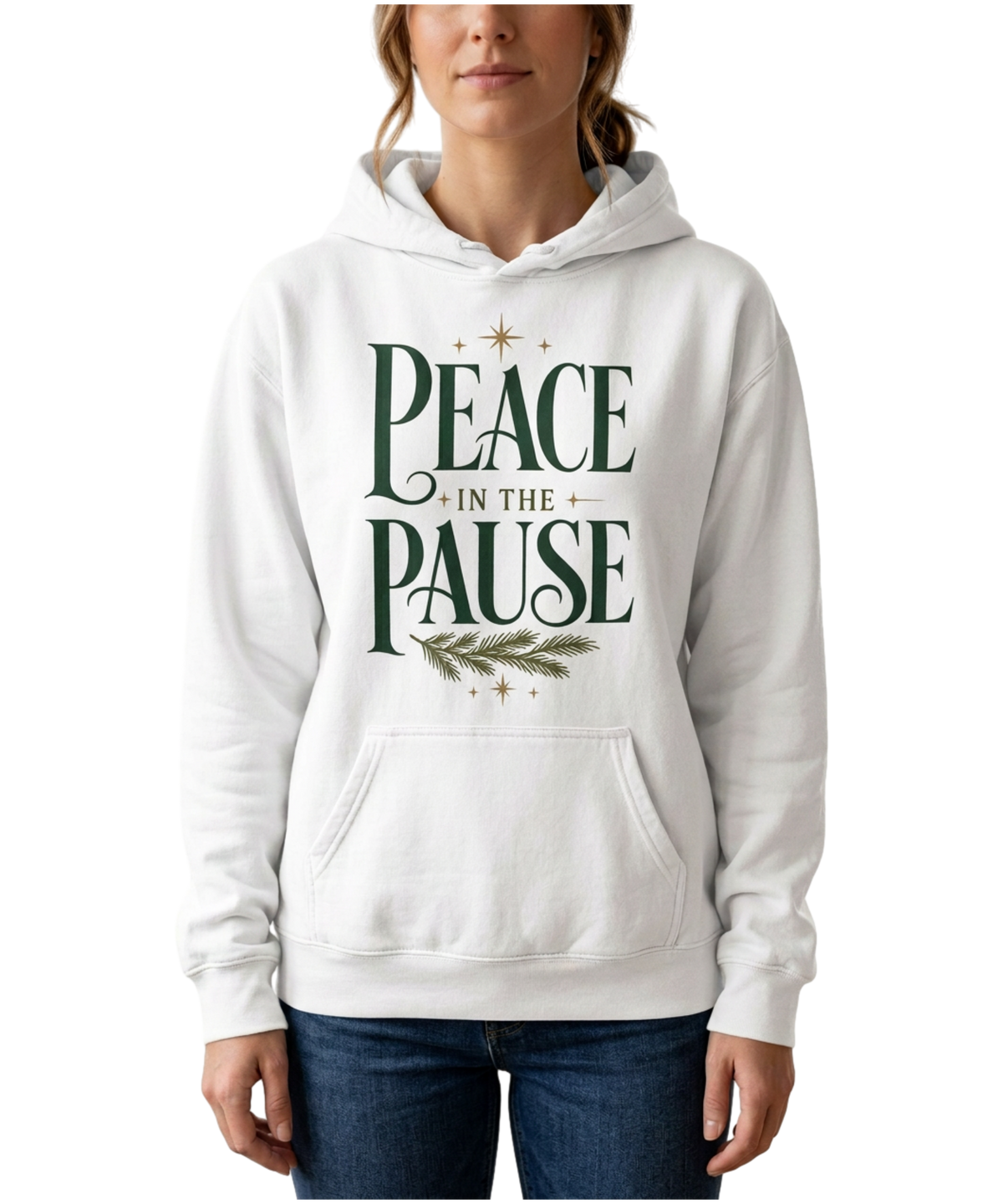 Model wearing a white Peace in the Pause hoodie with forest-green lettering, an evergreen sprig, and gold stars, paired with jeans against a plain white background.