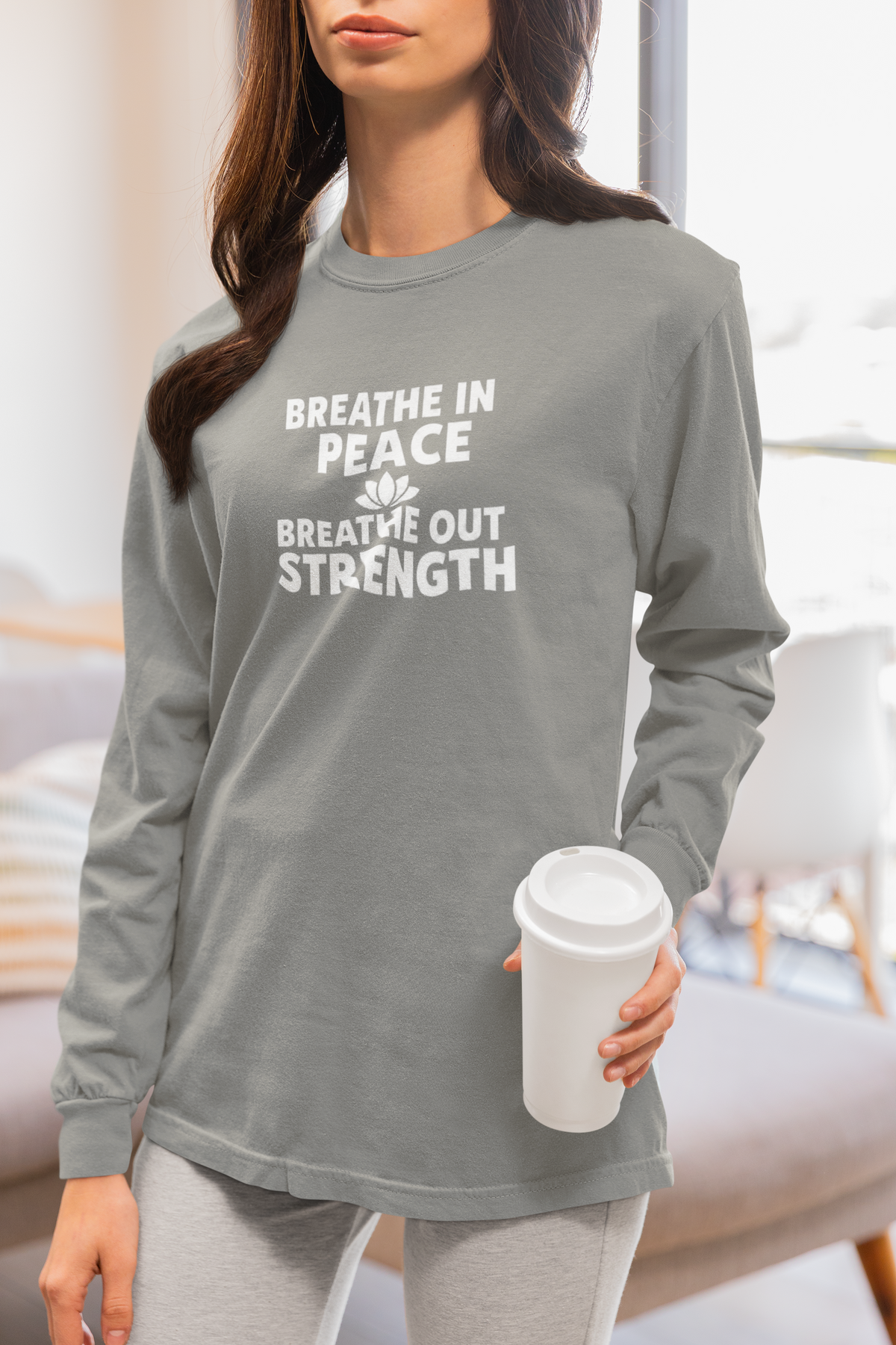 A woman wearing a heather gray long-sleeve t-shirt with the white printed text 'BREATHE IN PEACE BREATHE OUT STRENGTH' and a small leaf graphic, holding a white coffee cup in a brightly lit room.