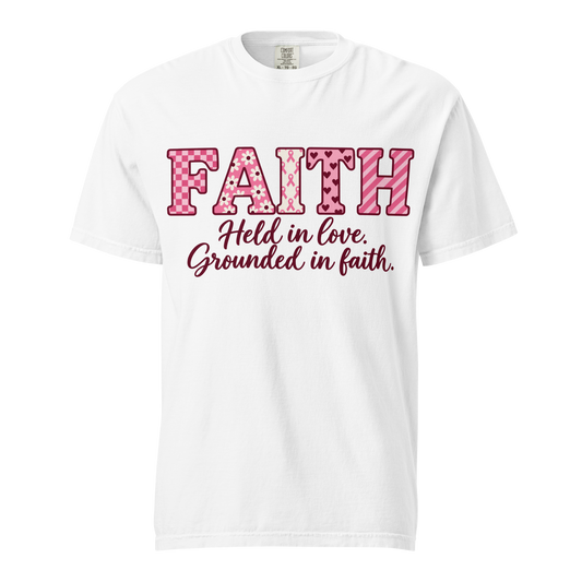 White T-shirt with pink patterned FAITH lettering, awareness ribbon motifs, and burgundy “Held in love. Grounded in faith.” script.
