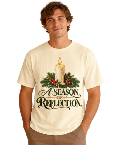 Man wearing an Ivory “A Season of Reflection” Christmas tee featuring a lit candle, evergreen branches, red berries, and green-and-gold lettering, paired with brown pants.