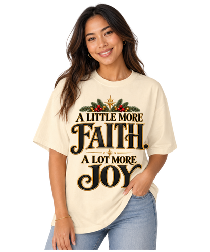 Woman wearing an Ivory “A Little More Faith, A Lot More Joy” Christmas tee with black-and-gold lettering, festive greenery, red berries, and star accents, paired with blue jeans.