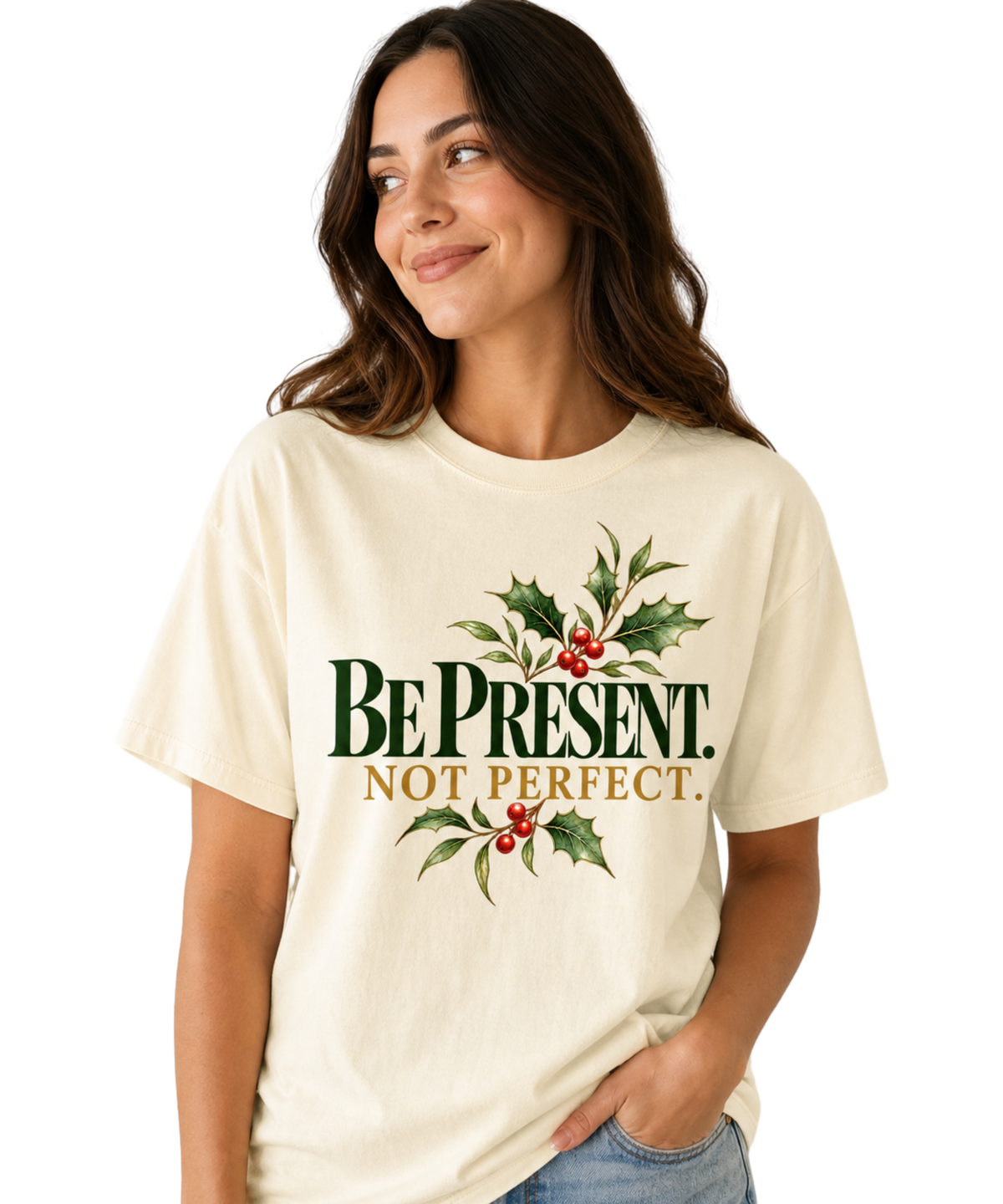 Woman wearing an Ivory “Be Present. Not Perfect.” T-shirt with green-and-gold lettering, holly leaves, and red berries, paired with blue jeans.