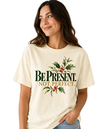 Woman wearing an Ivory “Be Present. Not Perfect.” T-shirt with green-and-gold lettering, holly leaves, and red berries, paired with blue jeans.