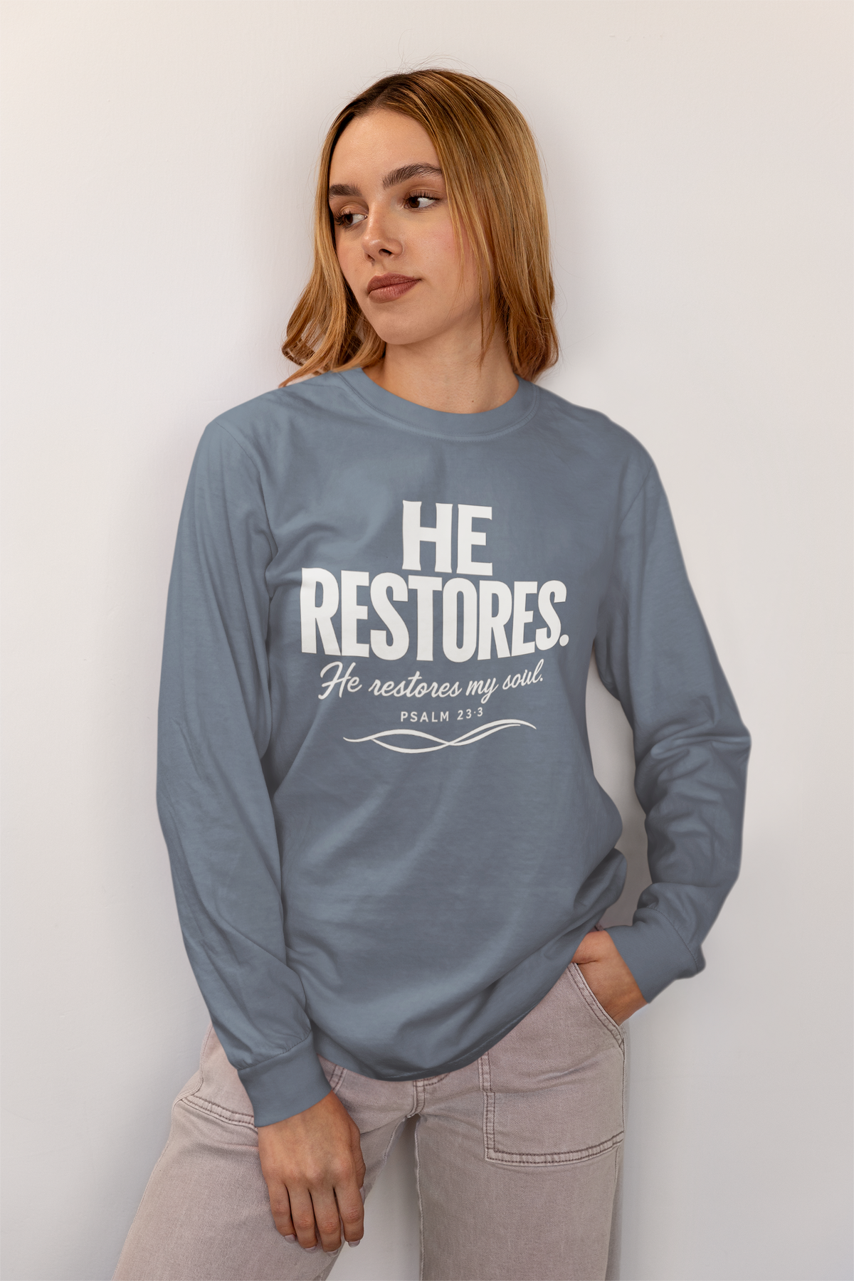 A woman wearing a slate blue long-sleeve crewneck sweatshirt with a white graphic print that reads 'HE RESTORES. He restores my soul PRAYS FOR US Psalm 37' paired with light gray jeans, standing against a plain off-white wall.