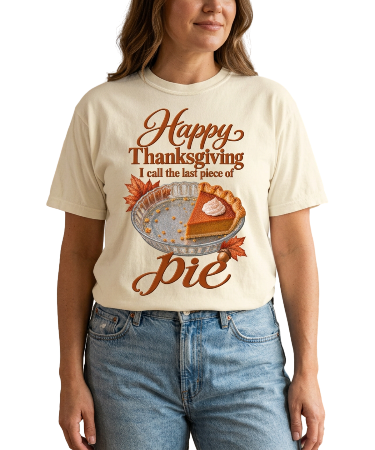 Woman wearing an ivory “I Call the Last Piece of Pie” tee featuring a pumpkin-pie slice and autumn leaves, paired with light blue jeans in a warm neutral interior.