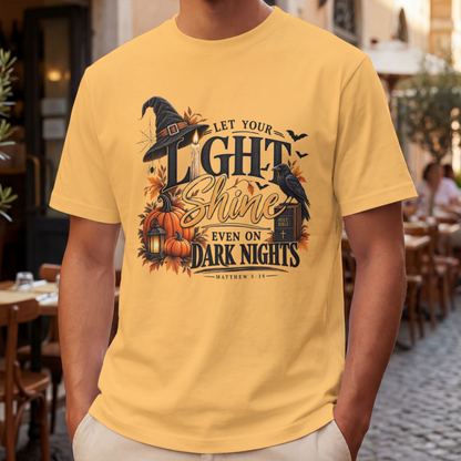 A man wearing a mustard graphic t-shirt with a Halloween-themed design that reads 'LET YOUR LIGHT SHINE EVEN ON DARK NIGHTS' with pumpkins, a witch hat, and a raven, set against a blurred outdoor cafe background.
