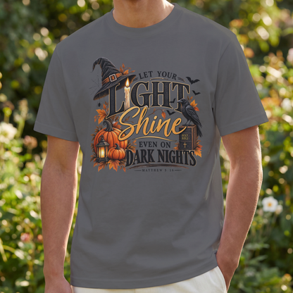A man wearing a pepper-color t-shirt with a Halloween graphic that reads 'LET YOUR LIGHT SHINE EVEN ON DARK NIGHTS' with pumpkins, a witch hat, and a raven, set against a blurred garden background.