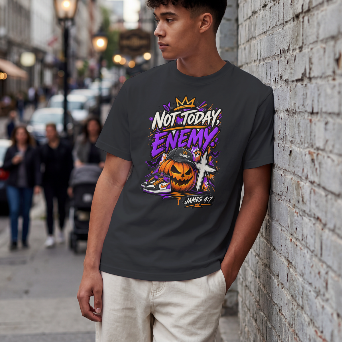 A man wearing a greyish black graphic t-shirt stands against a white brick wall on a city street. The t-shirt features a colorful graphic with a crown, the text 'NOT TODAY, ENEMY!' in purple and white, a jack-o'-lantern, a cross, and the text 'JAMES 4:7'.