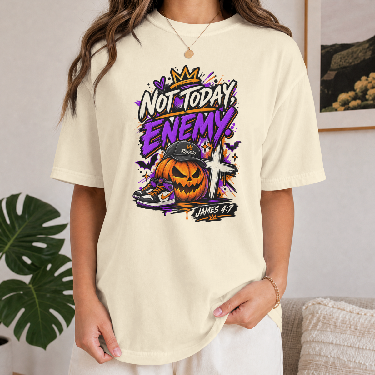 A woman wearing an ivory-colored graphic t-shirt with a Halloween-themed design that reads 'NOT TODAY, ENEMY.' and features a jack-o'-lantern, cross, and sneaker.