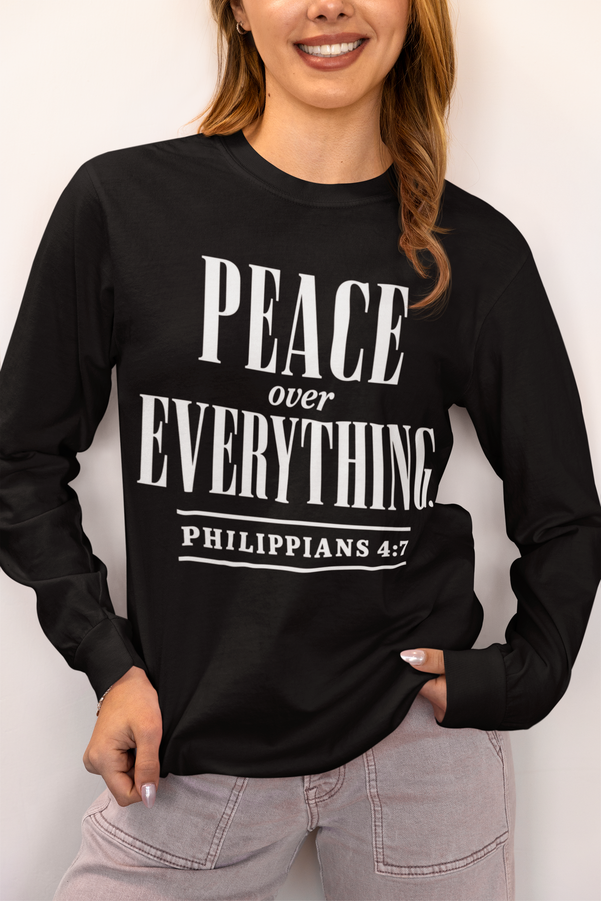 A woman wearing a black long-sleeve graphic shirt with the text 'PEACE over EVERYTHING PHILIPPIANS 4:7' printed in bold white letters, paired with light gray jeans against a plain off-white background.