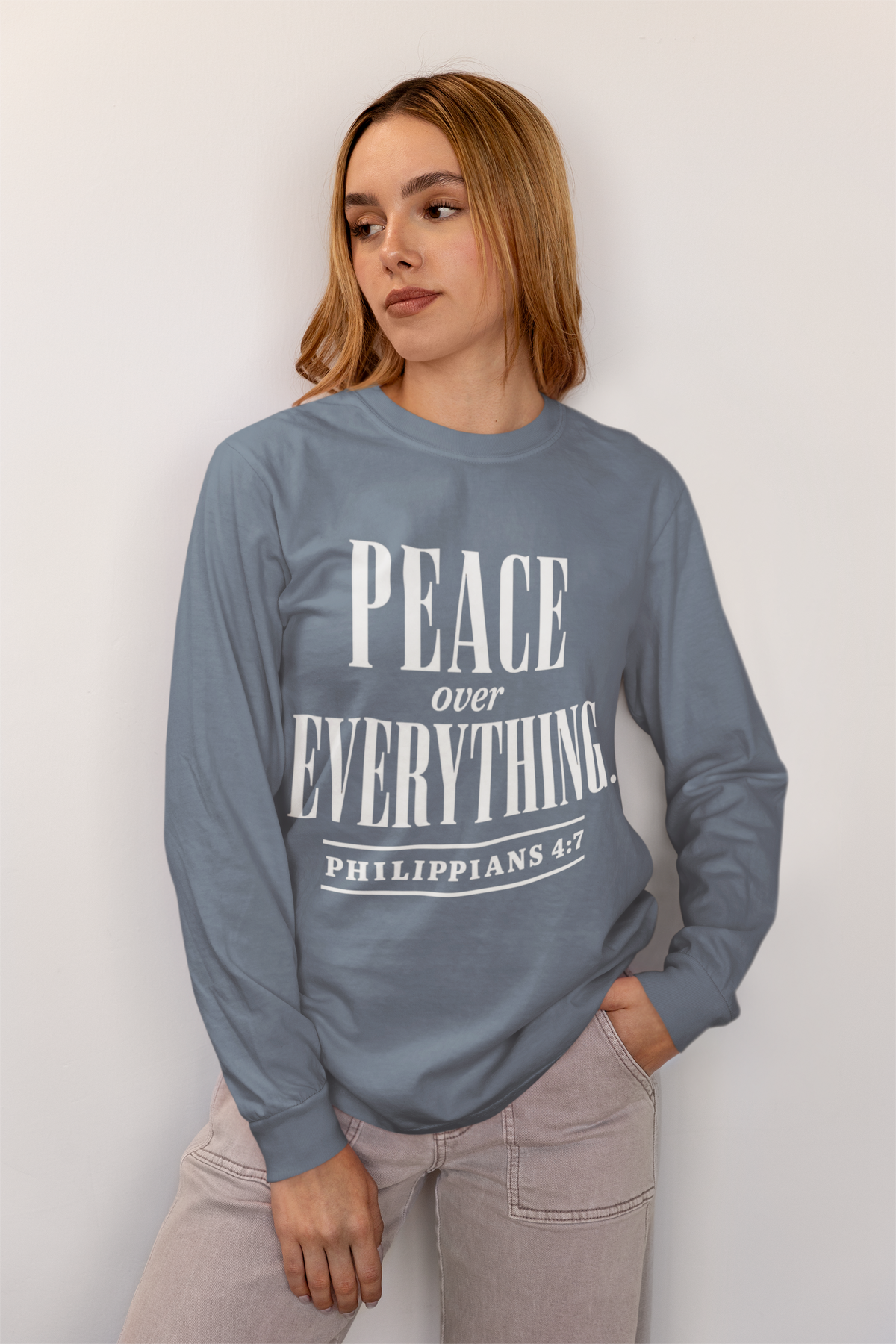 A woman wearing a slate blue long-sleeve crewneck sweatshirt with 'PEACE over EVERYTHING. PHILIPPIANS 4:7' printed in white on the front, paired with light gray jeans, set against a plain off-white wall.