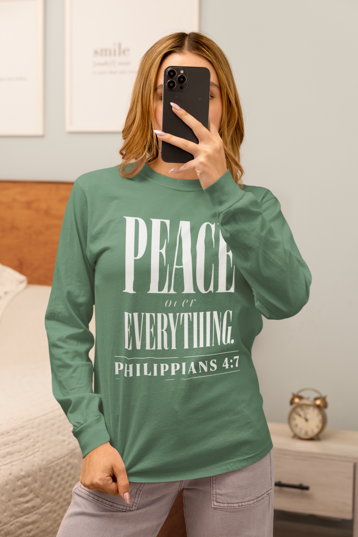 A woman taking a mirror selfie in a bedroom wearing a sage green long-sleeve t-shirt with a white graphic print that reads 'PEACE over EVERYTHING. PHILIPPIANS 4:7'.