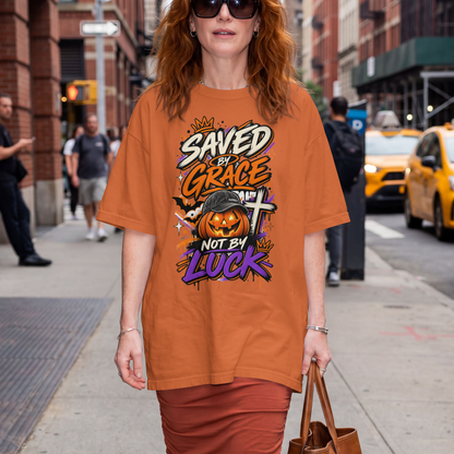 A woman stands on a city sidewalk wearing an oversized yam-colored graphic T-shirt with a Halloween-themed print reading 'Saved by Grace Not by Luck' and a jack-o'-lantern, plus matching burnt orange skirt.