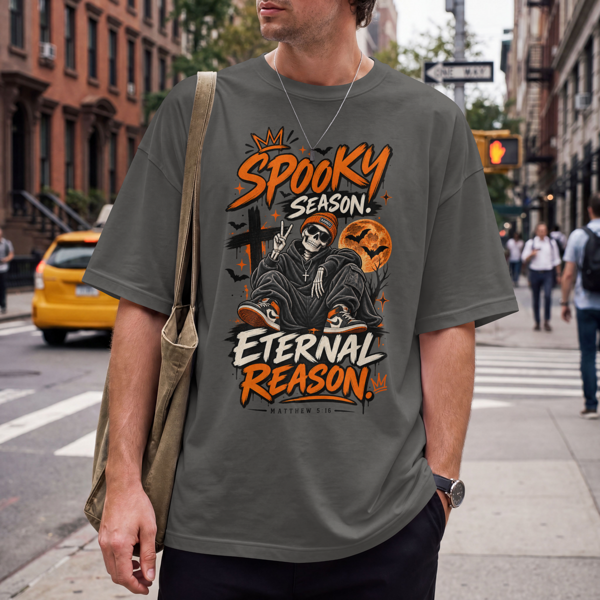 A man wearing a pepper graphic t-shirt with a Halloween design that reads 'Spooky Season Eternal Reason'. He is carrying a tan shoulder bag on a city street with brick buildings and a yellow cab in the background.