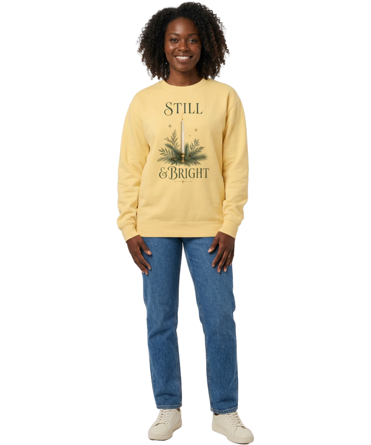 Female model wearing a Butter yellow Still & Bright Christmas sweatshirt with candle and evergreen artwork, blue jeans, and cream sneakers.