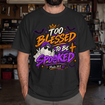 A man wearing a black graphic t-shirt with a colorful Halloween-themed print that reads 'Too Blessed to Be Spooked Psalm 34:4' in orange, purple, and white, set against a blurred garage background.