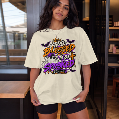 A woman wearing an ivory-colored graphic t-shirt with a Halloween-themed design that reads 'Too Blessed to Be Spooked' in orange, purple, and black lettering, paired with black shorts, standing in a modern indoor setting.