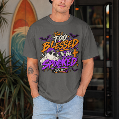 A man wearing a pepper-colored graphic t-shirt with a 'Too Blessed to Be Spooked' Halloween design, standing in front of a blurred background with a surfboard and plants.