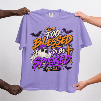 A violet graphic t-shirt held up by four hands against a light gray background. The shirt features a colorful Halloween-themed graphic with the text 'TOO BLESSED TO BE SPOOKED' and 'Psalm 34:14' with cartoon ghosts, bats, and a purple witch hat.