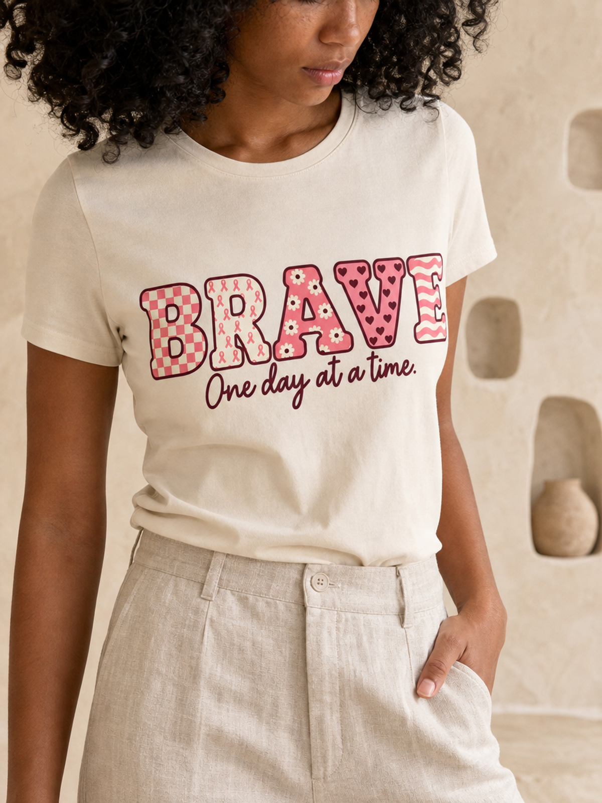 Woman wearing an ivory BRAVE graphic T-shirt tucked into light trousers, with pink patterned lettering and “One day at a time” script.
