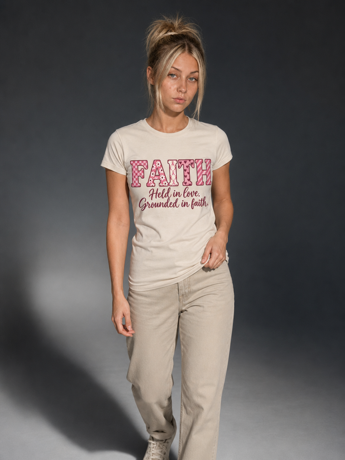 A woman modeling a light beige graphic t-shirt with pink and black text that reads 'FAITH Hold in love Grounded in faith' against a dark gray studio background.