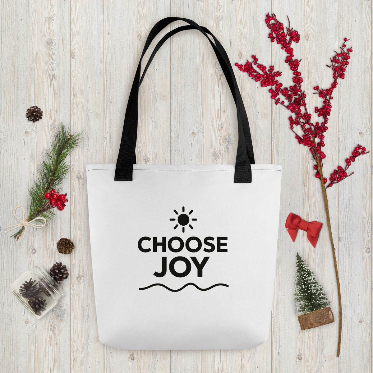 White Choose Joy tote with black handles on pale wood, styled with evergreen, red berries, and holiday decorations.