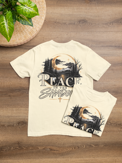Two ivory-colored t-shirts displayed on a wooden surface, showing the back design of a larger shirt and a smaller matching shirt in front. The back shirt features a graphic of a full moon over pine trees with the text 'PEACE IN THE SHADOW'.
