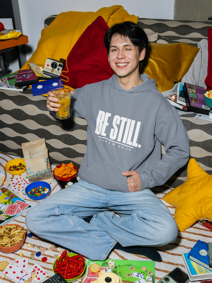 A smiling young man sits cross-legged on a patterned couch surrounded by snacks, cards, and toys in a cozy room setting. He is wearing a gray hoodie with the text 'BE STILL.