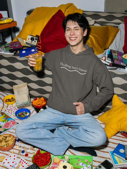 Person wearing a pepper (dark grey) hoodie with 'Healing isn't linear' text, sitting on a bed with snacks and games.