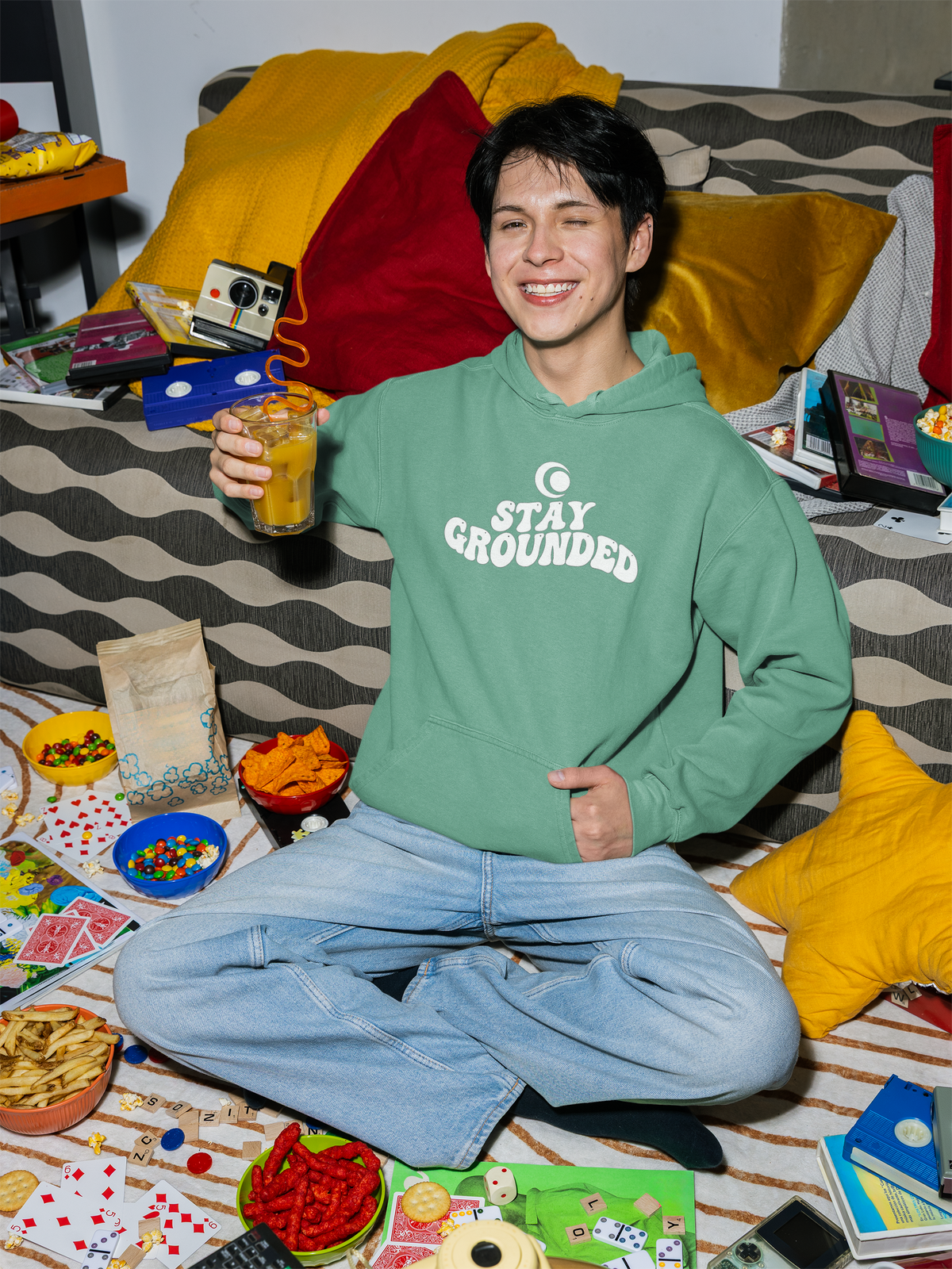 Person wearing a seafoam green hoodie with 'Stay Grounded' text, sitting on a bed with snacks and games around.