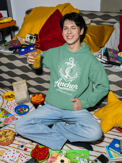 Person wearing a seafoam (green) hoodie with an anchor design, sitting on a bed with snacks and games around.