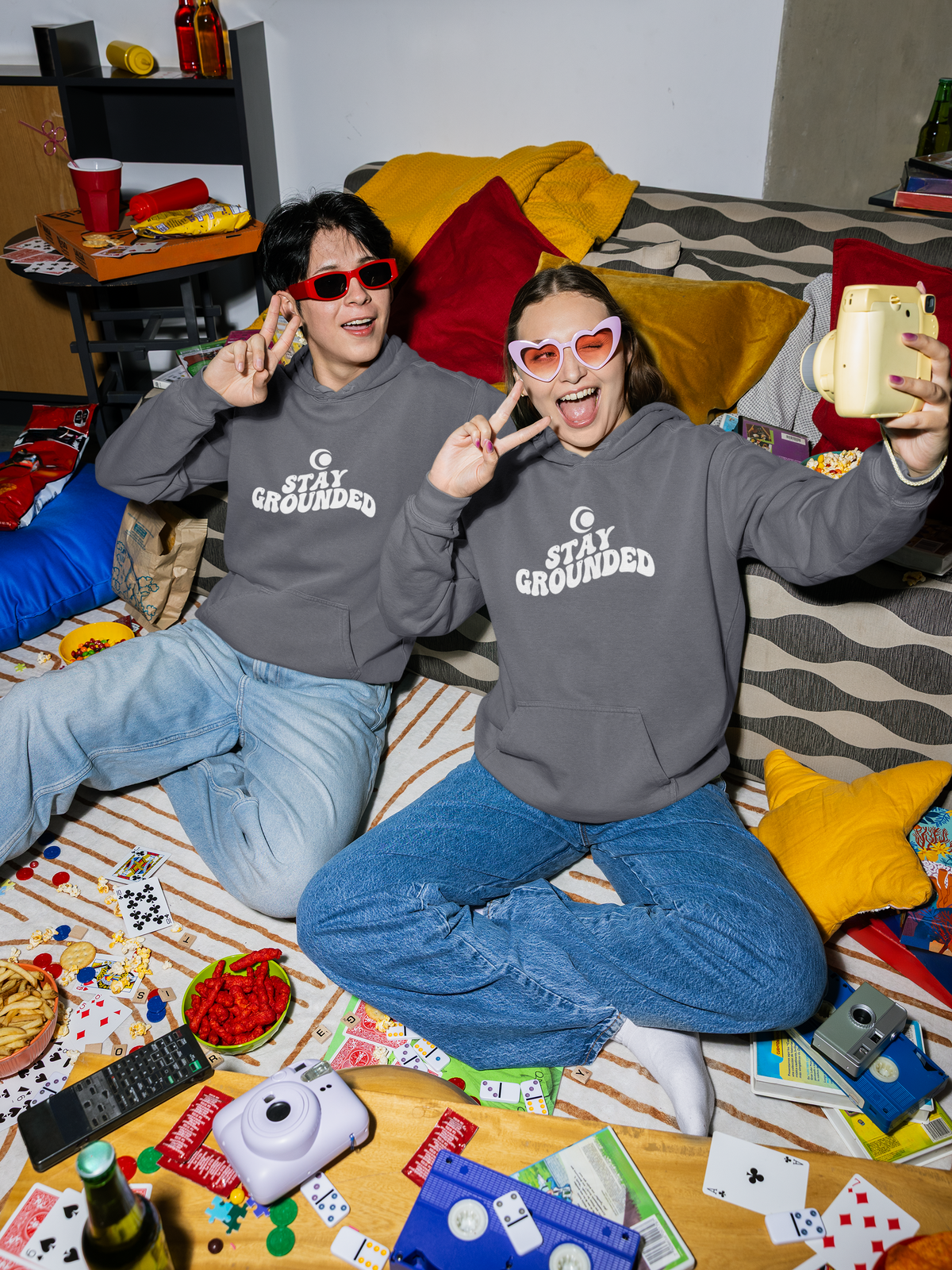 Two people wearing 'Stay Grounded' hoodies sitting on a couch with a game board and snacks around.
