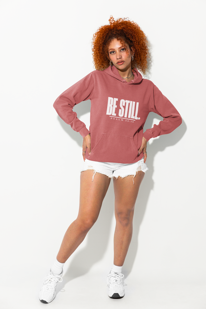A model with curly red hair poses in a dusty rose pink hoodie with 'BE STILL' printed in white on the chest, paired with white denim shorts and white sneakers against a white studio background.