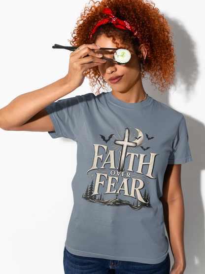 A model wearing a blue jean garment-dyed t-shirt featuring the Faith Over Fear cross graphic design with bats and crescent moon, in a casual lifestyle setting.