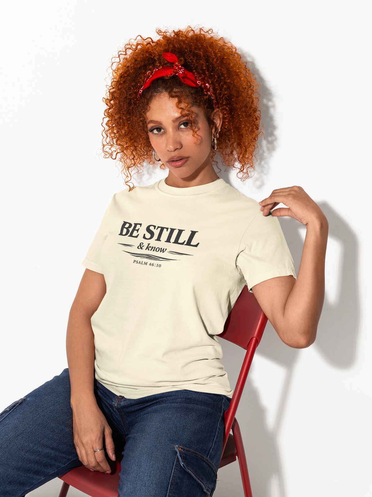 Woman wearing a 'Be Still & Know' t-shirt sitting on a red chair against a white background