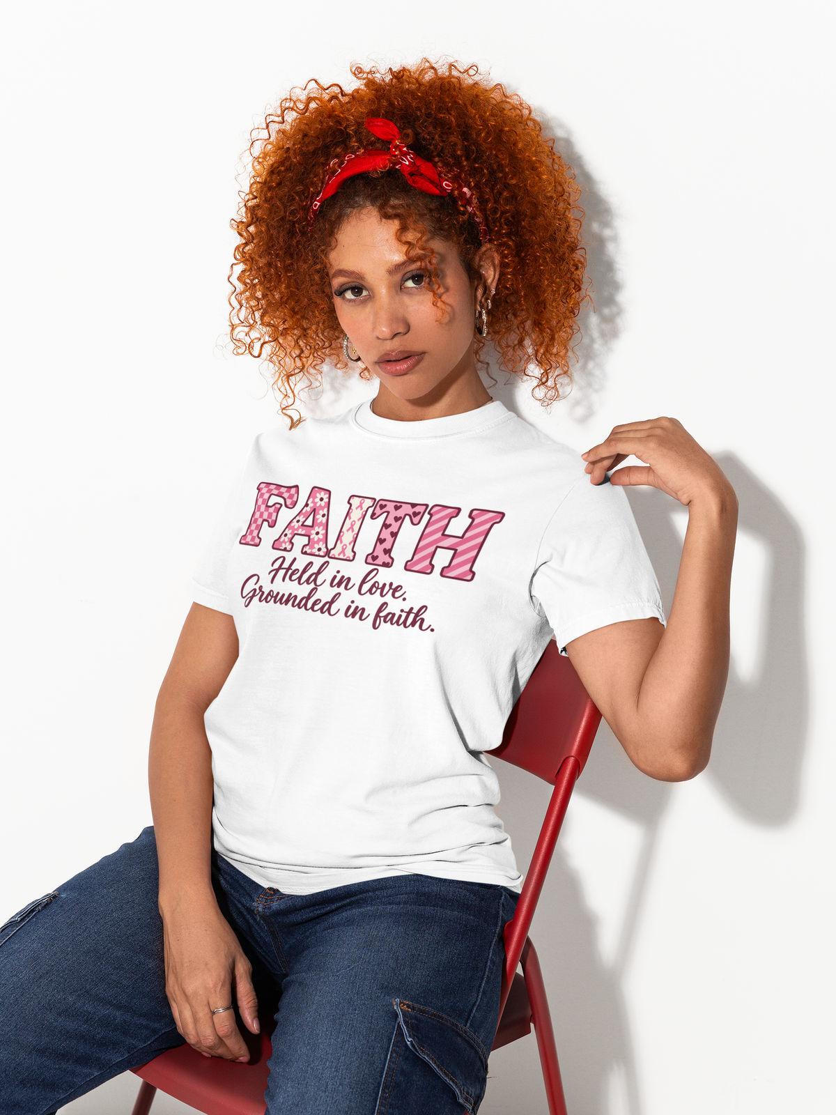 A woman with curly red hair sits on a red chair wearing a white graphic T-shirt with pink text reading 'FAITH Holds in love, Grounded in faith.' and blue jeans against a plain white background.
