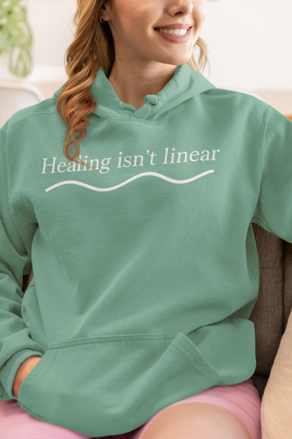 Person wearing a seafoam green hoodie with 'Healing isn't linear' text.
