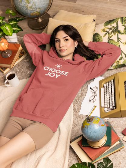 Woman wearing a crimson (pinkish) hoodie with 'CHOOSE JOY' text, lying on a bed in a cozy room.
