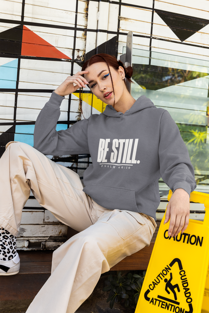 A woman sits on a bench wearing a gray pullover hoodie with 'BE STILL.' printed in white across the chest, paired with beige pants and sneakers.