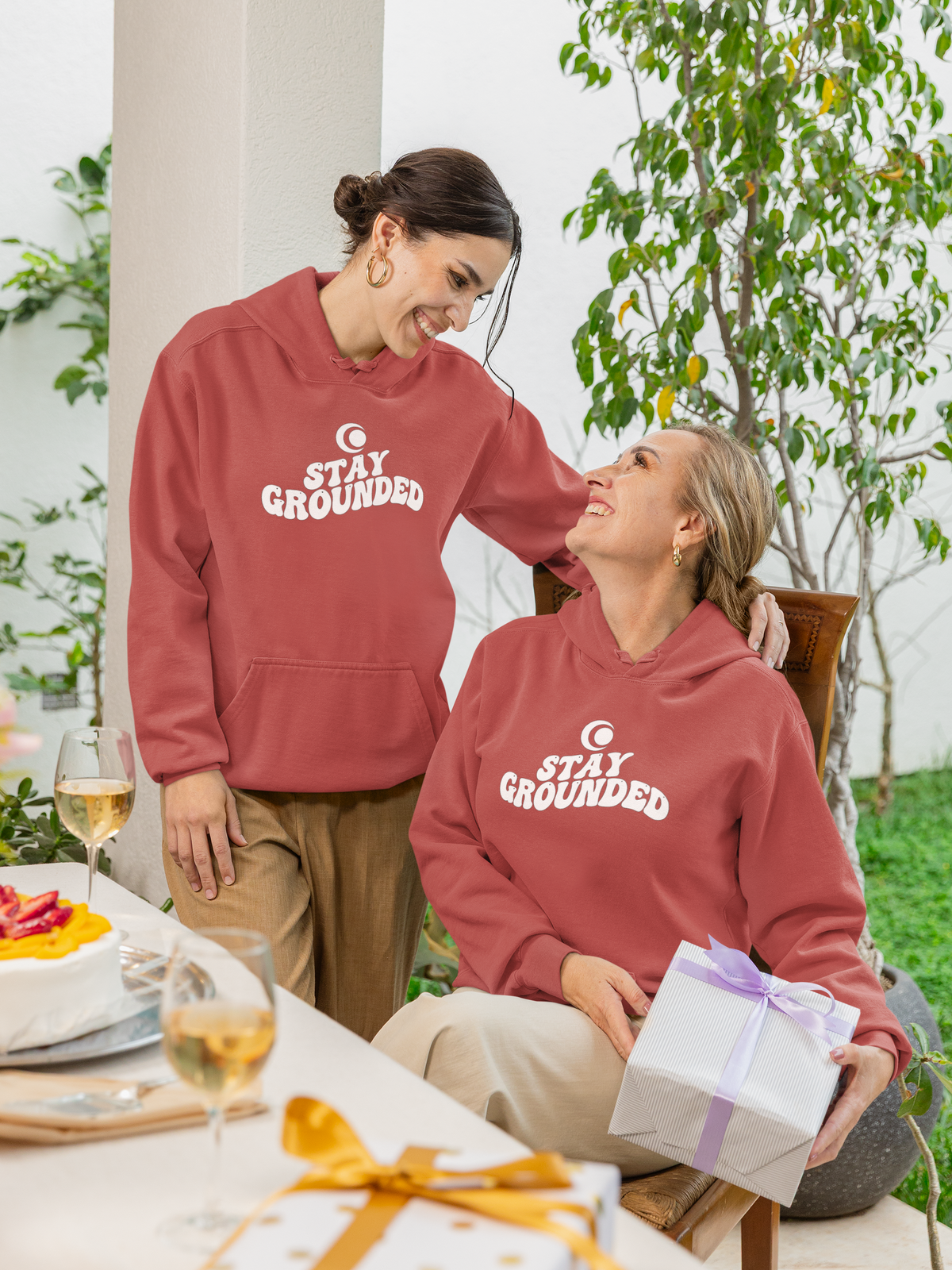 Two people wearing 'Stay Grounded' hoodies outdoors with a gift and wine glasses on a table.