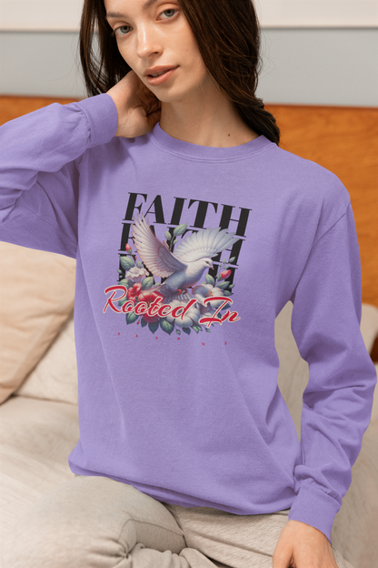 A woman wearing a lavender long-sleeve graphic sweatshirt with a dove and floral design and the words 'FAITH Rooted In', sitting on a couch in a bedroom setting.