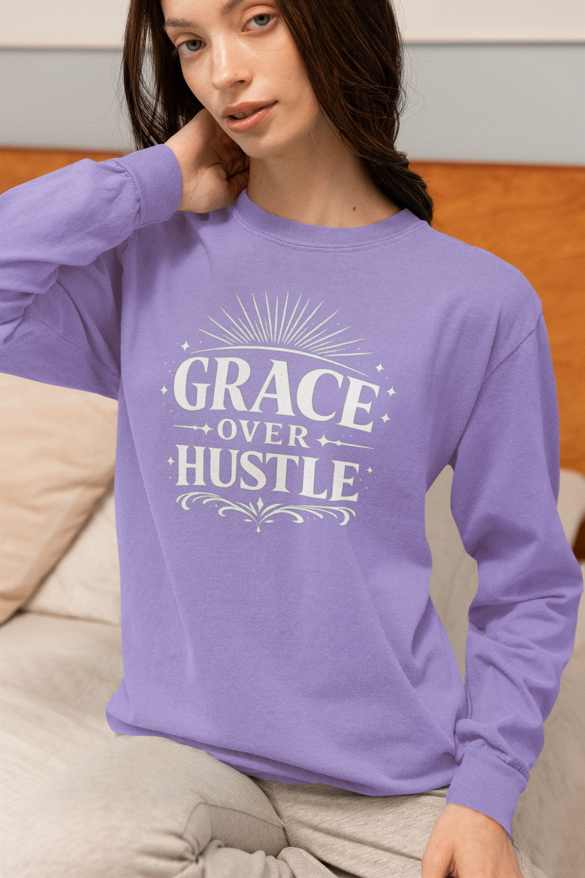 A woman wearing a lavender long-sleeve crewneck sweatshirt with a white graphic print that reads 'GRACE OVER HUSTLE' on the front, sitting on a beige couch.