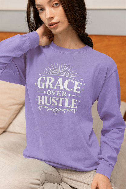 A woman wearing a lavender long-sleeve crewneck sweatshirt with a white graphic print that reads 'GRACE OVER HUSTLE' on the front, sitting on a beige couch.