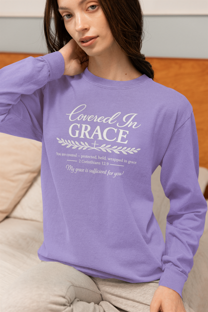 A woman wearing a lavender long-sleeve crewneck sweatshirt with white printed text reading 'BE STILL & know PSALM 46:10' sits on a beige couch in front of a wooden headboard.