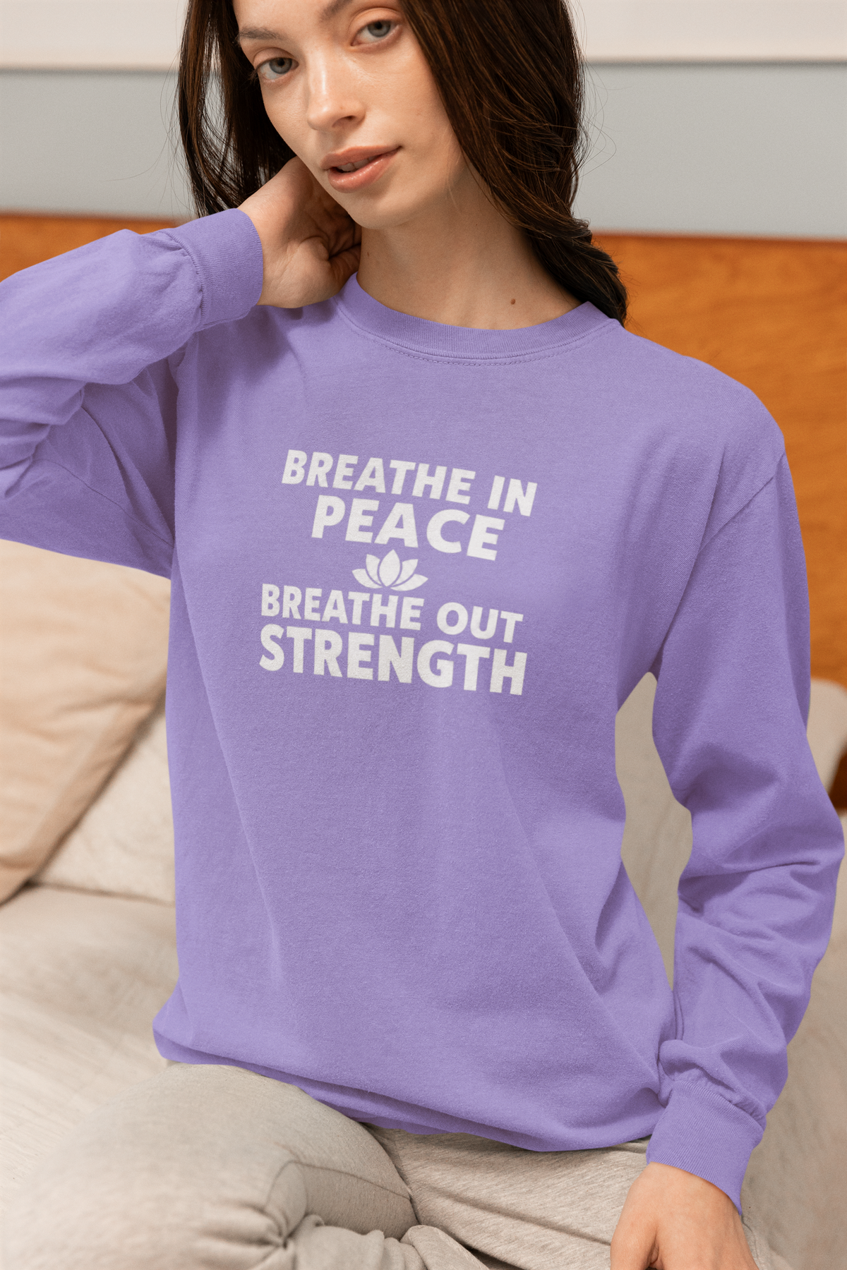 A woman wearing a lavender long-sleeve crewneck sweatshirt with the white printed text 'BREATHE IN PEACE BREATHE OUT STRENGTH' and a small lotus flower graphic, sitting on a beige couch.