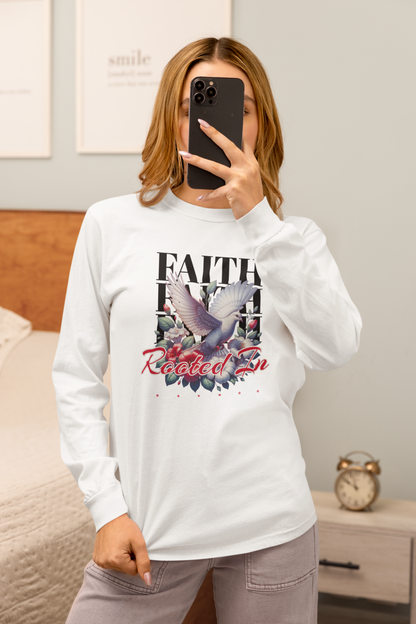 A woman takes a mirror selfie in a bedroom while wearing a white long-sleeve graphic t-shirt with a dove and floral design, paired with gray pants.