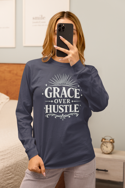 A woman takes a mirror selfie in a bedroom while wearing a navy blue long-sleeve t-shirt with a white graphic print that reads 'GRACE OVER HUSTLE'.