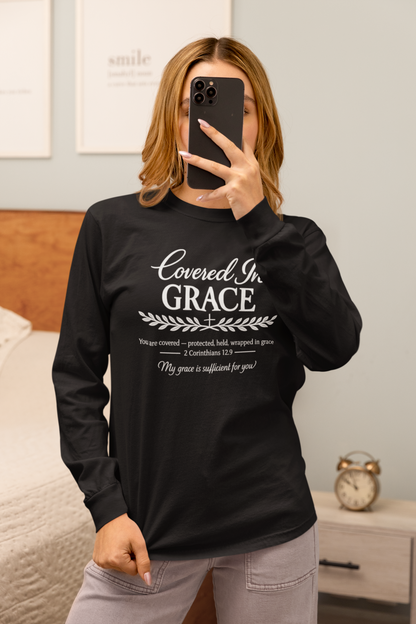 A woman takes a mirror selfie in a bedroom while wearing a black long-sleeve graphic t-shirt with white text that reads 'Covered in GRACE' and a Bible verse from 2 Corinthians 1:3, paired with light gray pants.