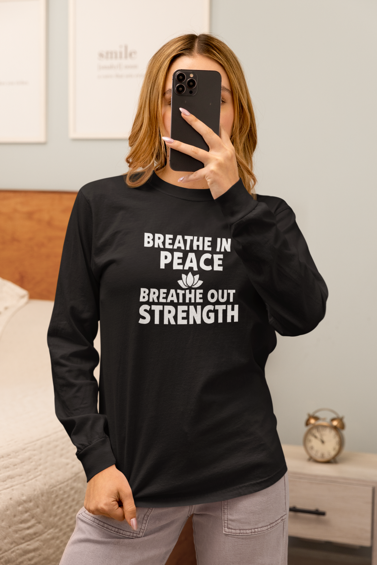 A woman takes a mirror selfie in a bedroom while wearing a black long-sleeve t-shirt with the white printed text 'BREATHE IN PEACE BREATHE OUT STRENGTH' and a small lotus flower graphic. She is wearing gray pants and holding a black smartphone.