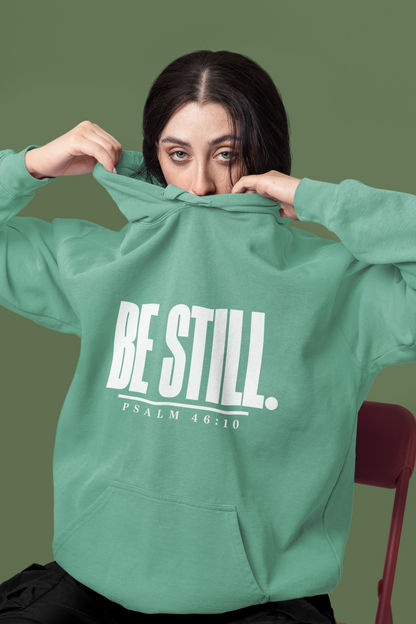A woman wearing a sage green hoodie with the text 'BE STILL. PSALM 48:10' printed in white on the front, holding the hood up over her face against a solid olive green background.